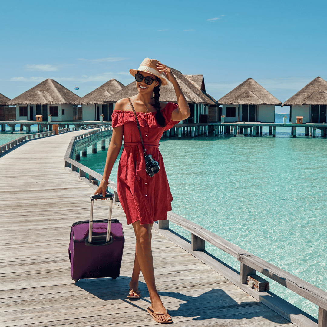 Woman in a red dress with a purple suitcase on a wooden pier with water bungalows in the background.
